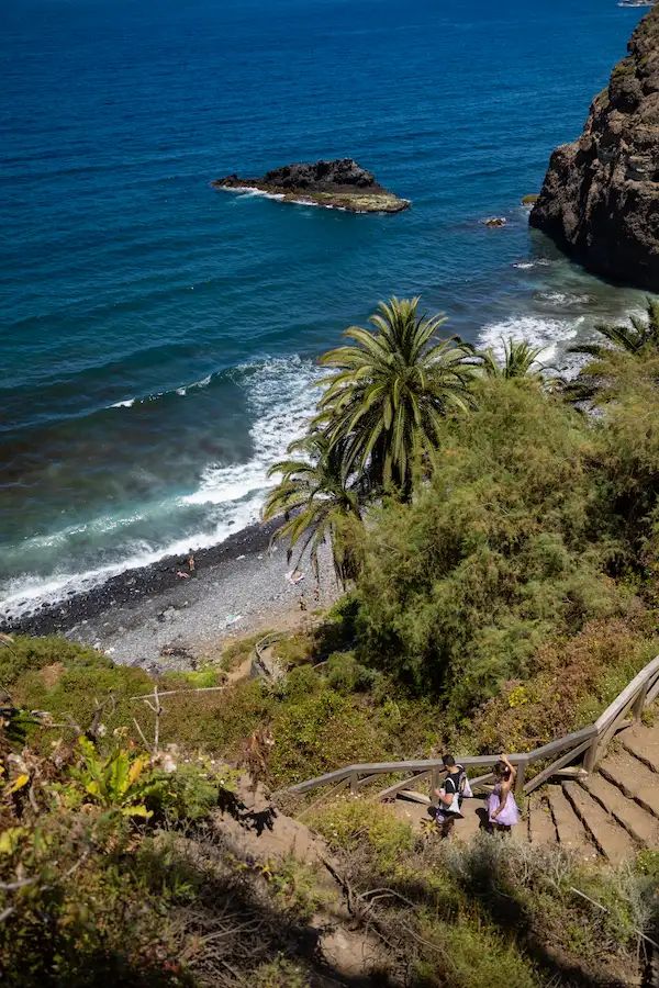 Vista desde el acantilado de la Rambla de Castro en Tenerife, con dos personas bajando por escaleras de madera hacia una playa de arena volcánica con palmeras y mar azul.